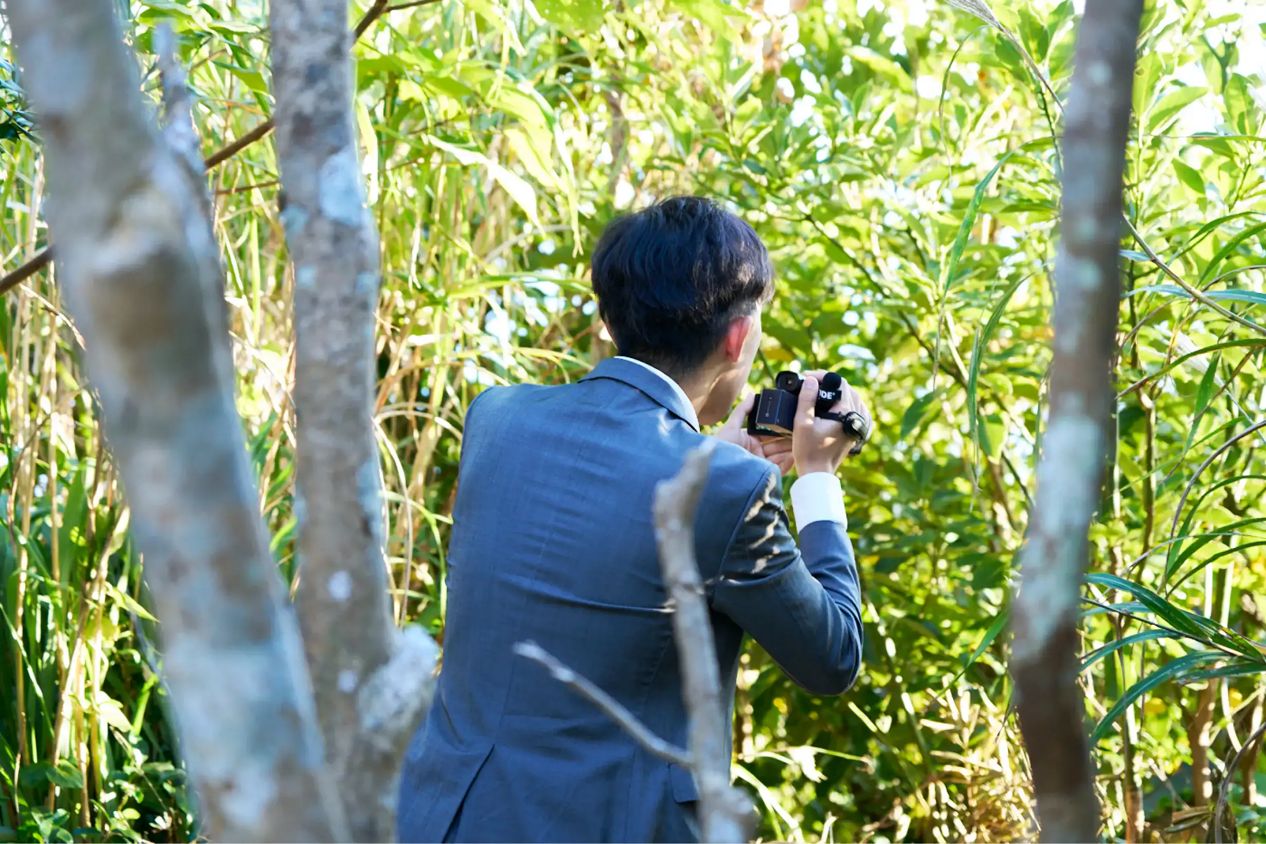 草木の中でカメラを構えるスーツさん
